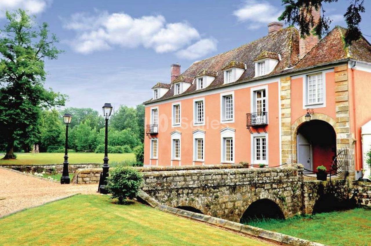 Manoir ancien en pierre avec façade rose, entouré de verdure, traversé par un petit pont en arc.