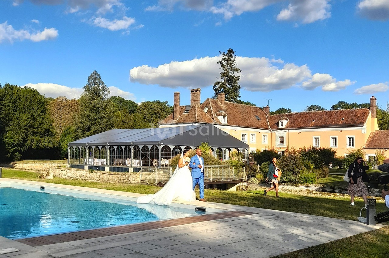 Un couple marche près d'une piscine dans un jardin, avec une grande maison et une tente en arrière-plan sous un ciel bleu.