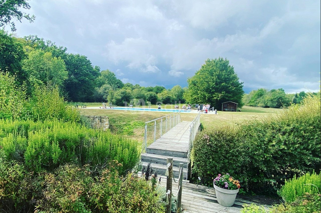 Passerelle en bois traversant un jardin verdoyant, menant à une prairie avec une piscine et des arbres en arrière-plan.