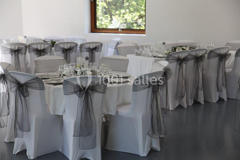 Salle de réception avec tables rondes dressées, nappes blanches et chaises décorées de nœuds en tissu noir.