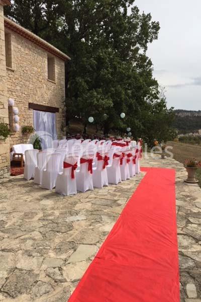 Chaises blanches avec nœuds rouges alignées en extérieur sur une terrasse en pierre, avec une allée rouge et un décor…