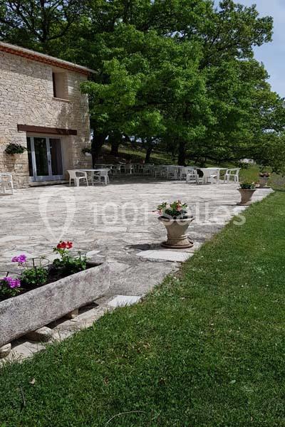 Terrasse en pierre avec tables et chaises blanches, bordée de verdure et d'arbres, près d'une maison en pierre.