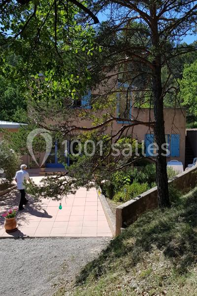 Maison en pierre avec volets bleus, entourée de végétation, une personne marche sur une terrasse ensoleillée.