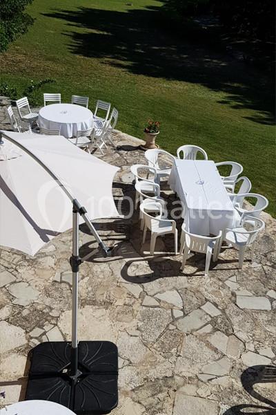 Tables et chaises blanches disposées sur une terrasse en pierre, avec un parasol et une pelouse en arrière-plan.