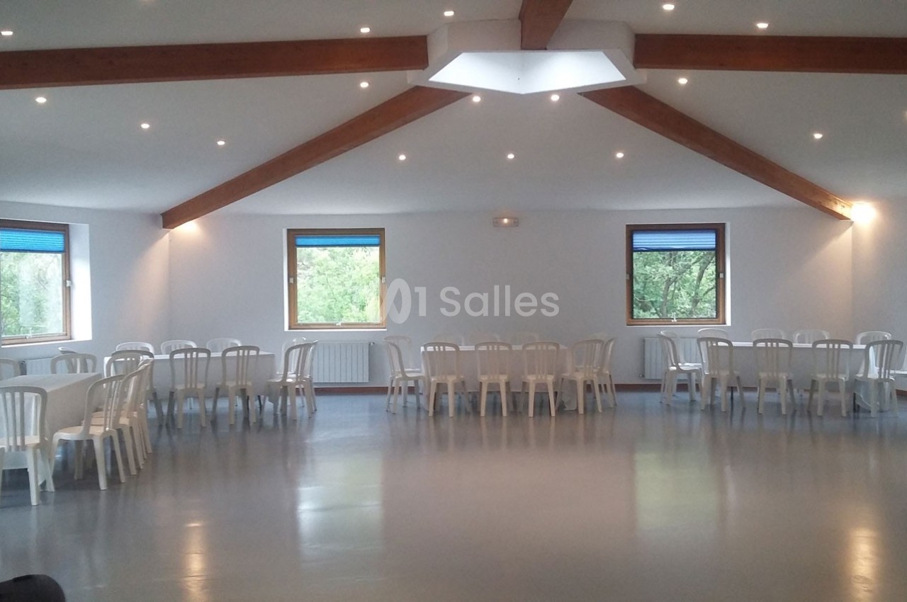 Salle lumineuse avec plafond en poutres apparentes, tables rondes et chaises blanches disposées le long des murs.