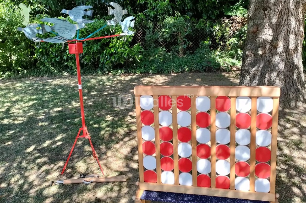 Un jeu de puissance 4 géant en bois placé sur une pelouse, à côté d'une girouette rouge sous des arbres.
