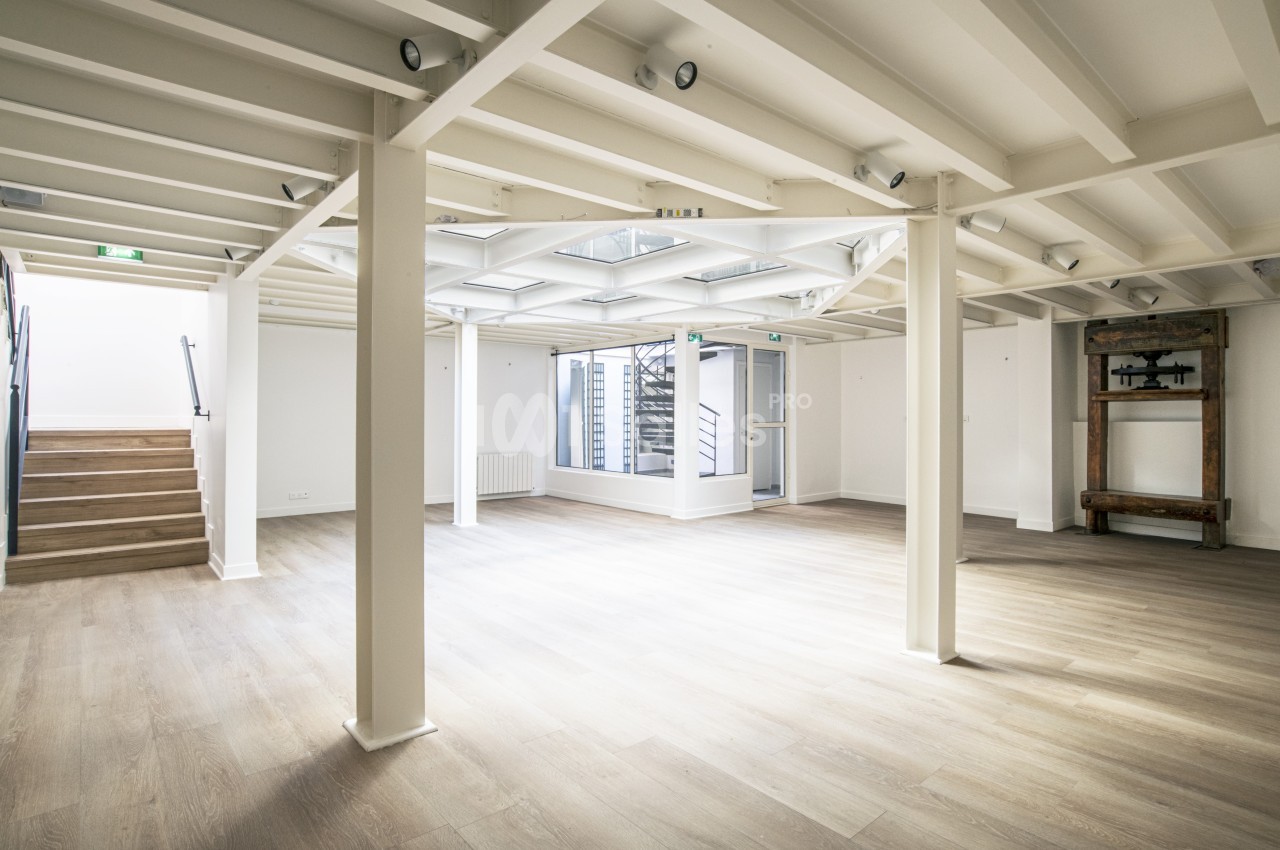 Salle lumineuse avec parquet clair, poutres apparentes blanches, escalier sur la gauche et grande verrière au plafond.