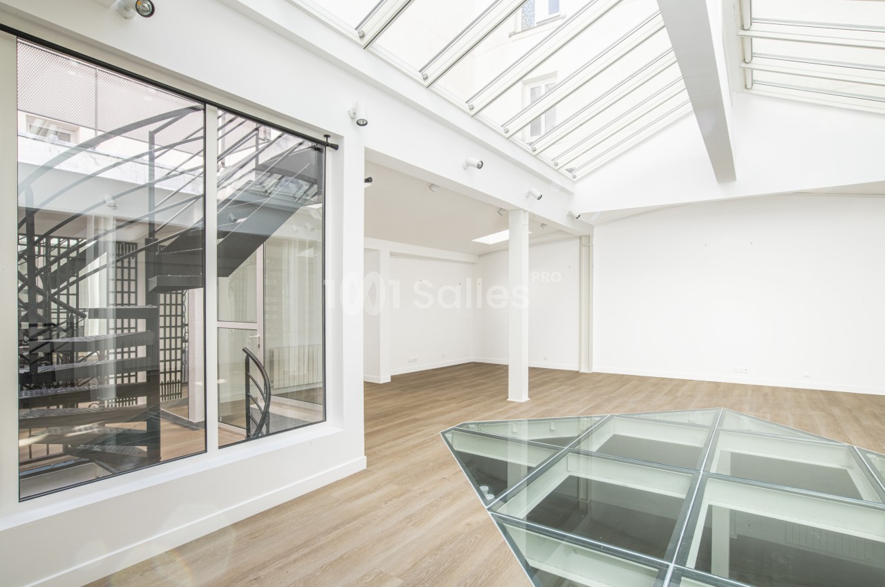 Intérieur lumineux avec verrière, escalier métallique en colimaçon et sol en parquet clair.
