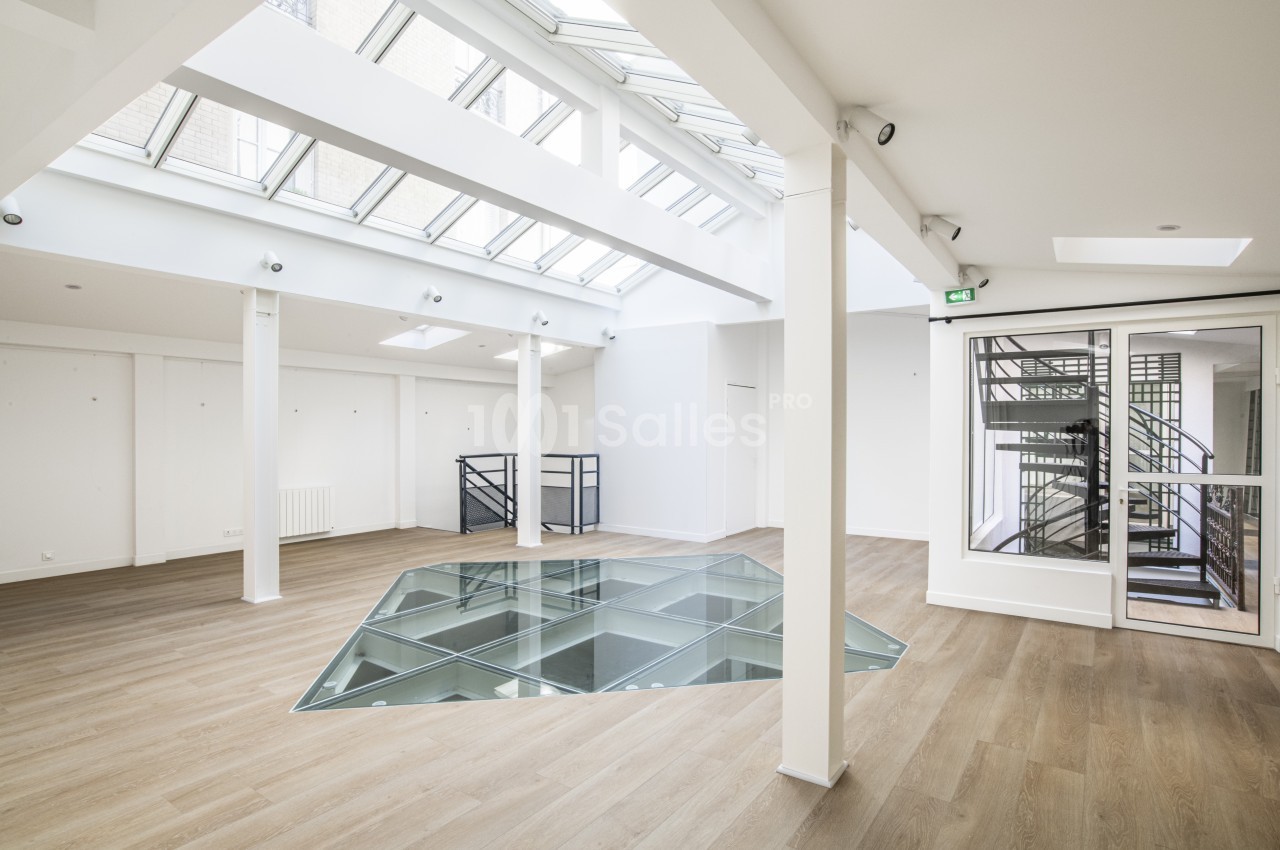Espace intérieur lumineux avec parquet, verrière au plafond, sol en verre et escalier visible derrière une porte vitrée.