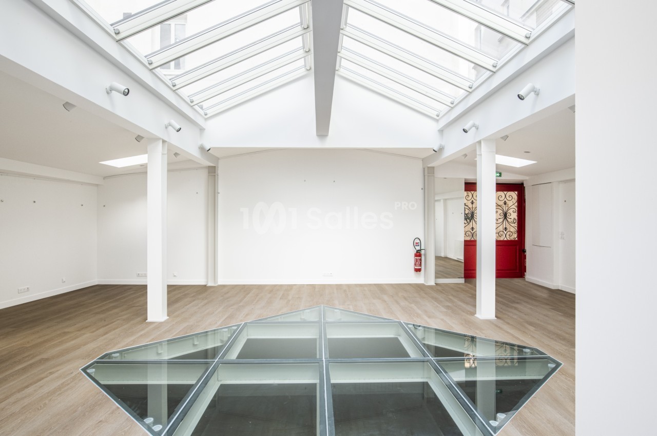 Intérieur lumineux avec verrière, sol en parquet clair et une trappe vitrée au centre, porte rouge au fond.