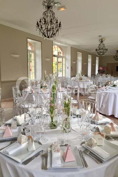 Salle de réception élégante avec tables rondes dressées, décorations florales et chaises transparentes.