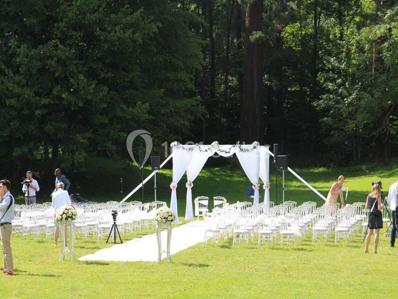Allée centrale bordée de chaises blanches menant à une arche décorée, installée dans un jardin verdoyant.