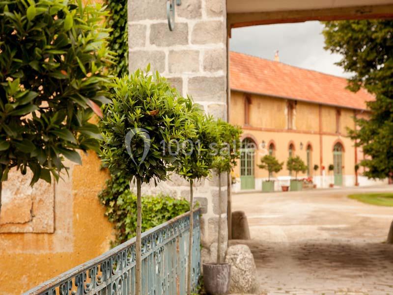 Allée bordée d'arbustes taillés en boule, menant à un bâtiment ancien avec des murs en pierre et des toits en tuiles.