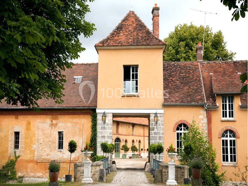Façade d'une maison ancienne avec toit en tuiles, porche central et cour intérieure visible à travers l'arche.