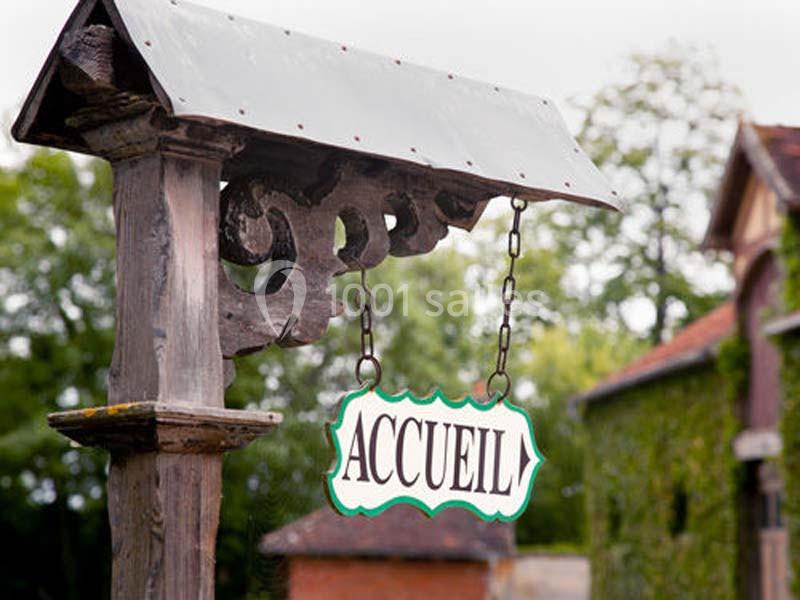 Enseigne en bois suspendue indiquant ’Accueil’, devant un bâtiment entouré de verdure.