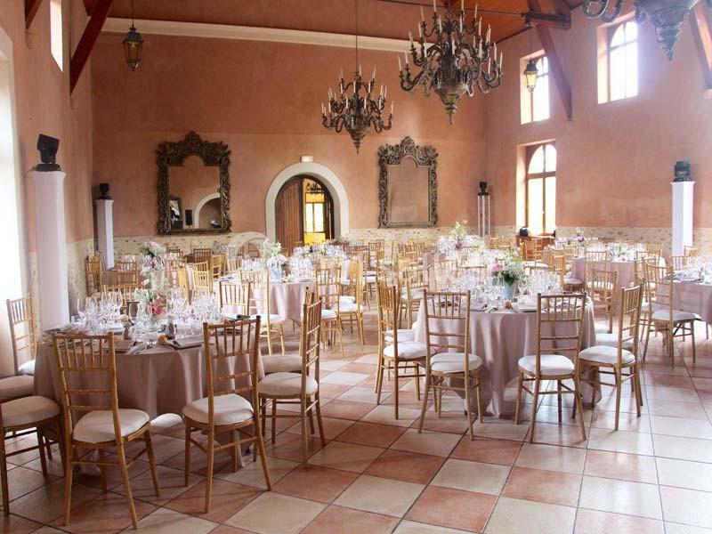 Salle de réception élégante avec tables rondes dressées, chaises dorées et lustres suspendus au plafond.