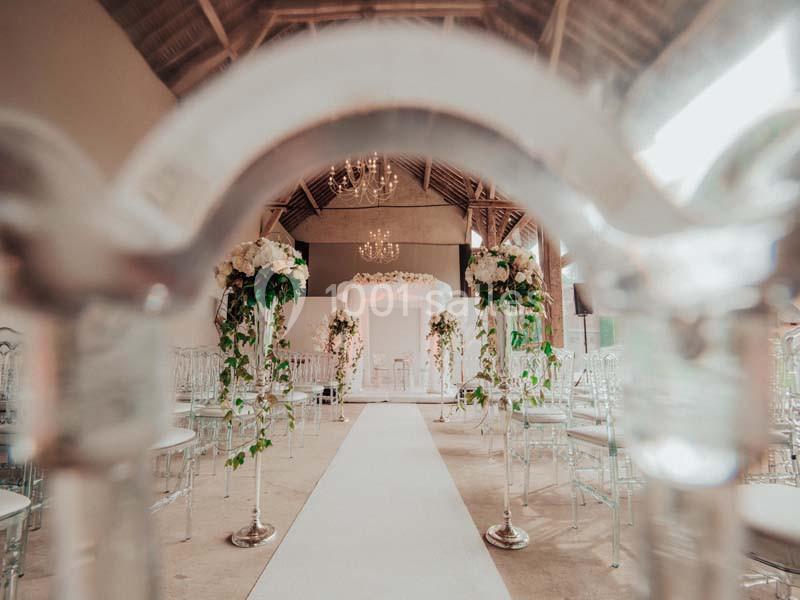 Allée centrale décorée de fleurs blanches et verdure, menant à un autel dans une salle de réception élégante.
