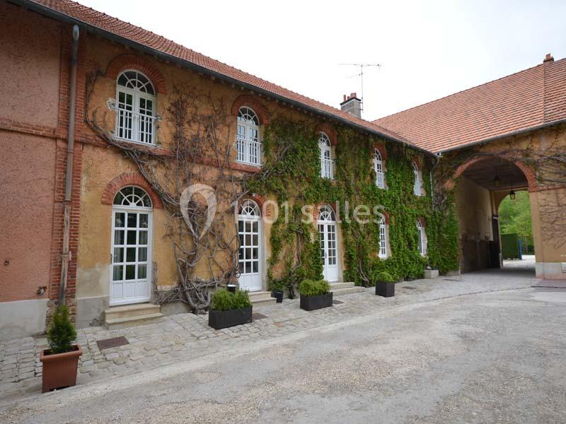 Façade d'un bâtiment ancien avec murs recouverts de vigne, grandes fenêtres blanches et cour pavée.