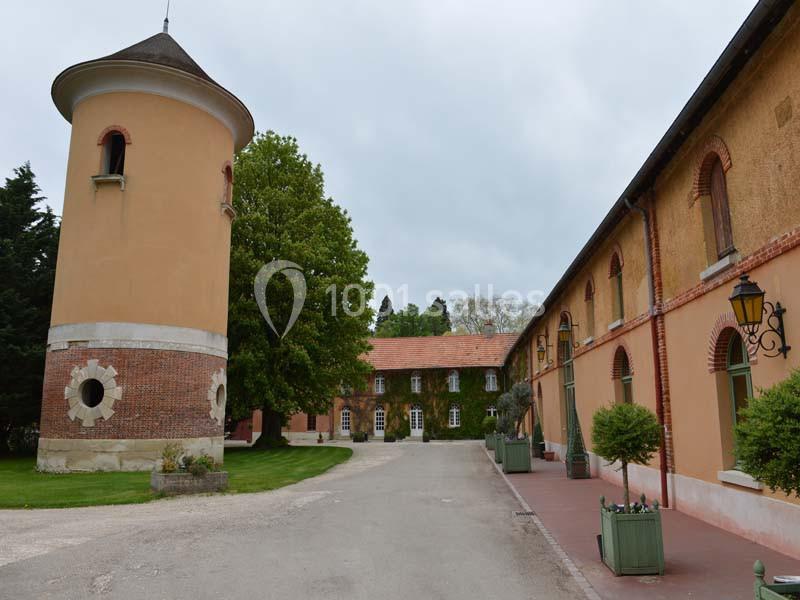 Bâtiment ancien avec une tour ronde, des façades en briques et un chemin bordé d'arbres et de lampadaires.