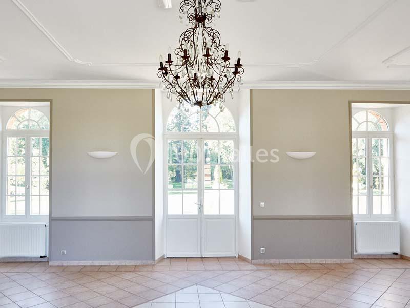 Salle lumineuse avec un lustre central, murs clairs, carrelage au sol et grandes fenêtres cintrées donnant sur un jardin.