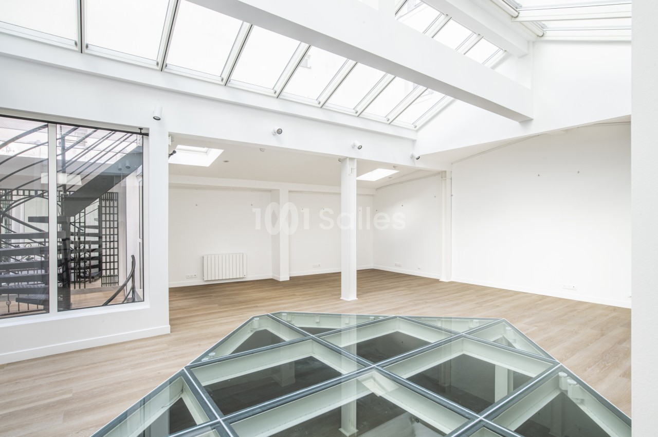 Espace intérieur lumineux avec parquet clair, verrière au plafond et sol vitré, escalier visible à travers une cloison…