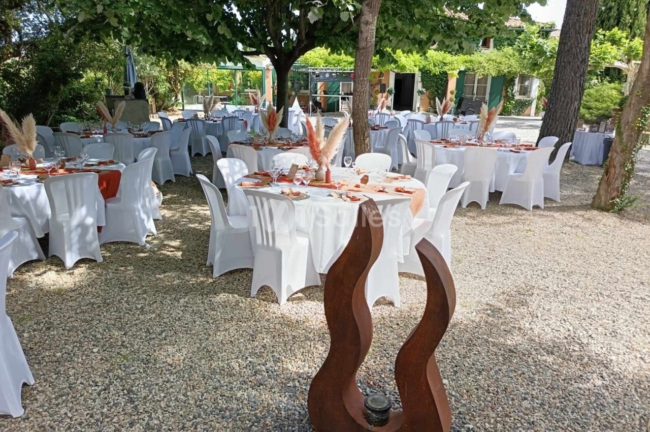 Tables décorées pour un événement en plein air, disposées sous des arbres, avec nappes blanches et ornements naturels.