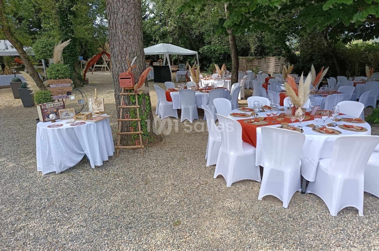 Tables rondes décorées pour un événement en extérieur, disposées sous des arbres avec nappes blanches et chemins de table…