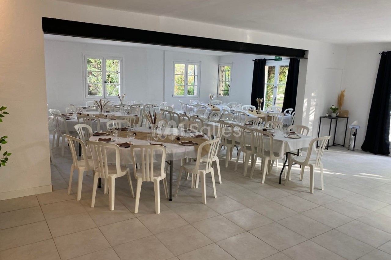 Salle lumineuse avec des tables rectangulaires dressées et entourées de chaises blanches, fenêtres donnant sur l'extérieur.