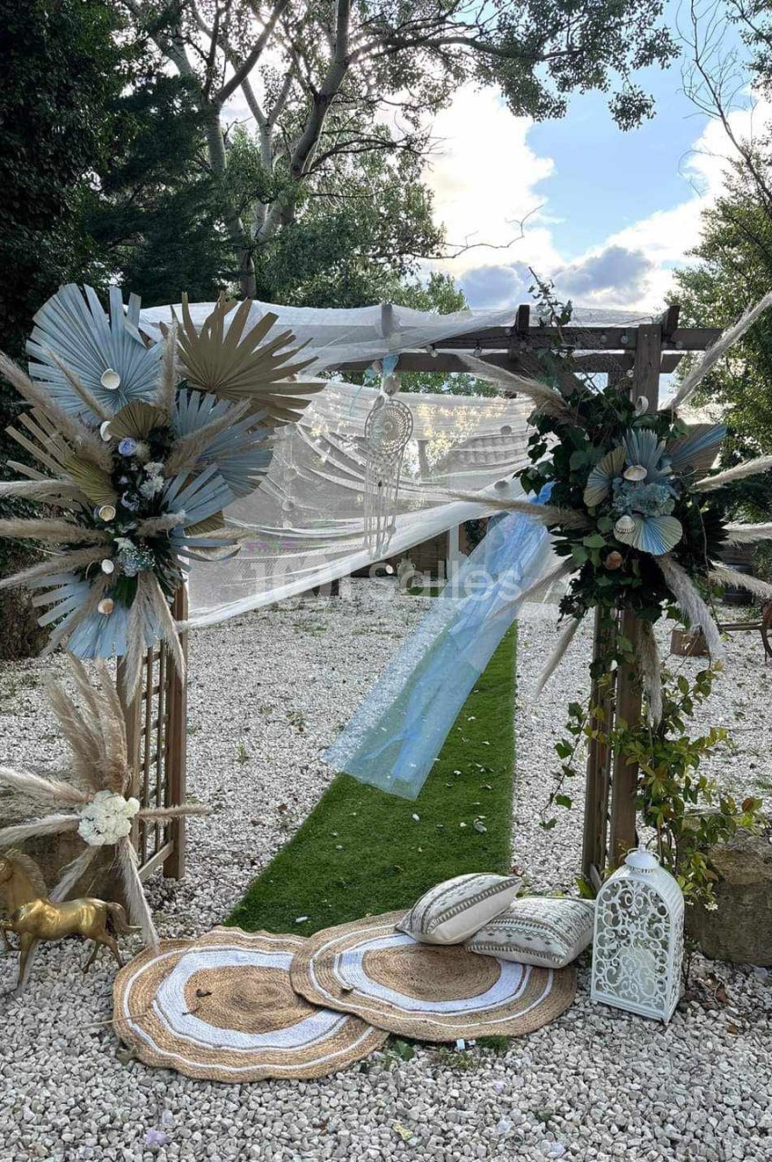 Arche décorée de fleurs séchées et voilages, entourée de tapis et lanternes, sur un chemin en gravier et gazon.