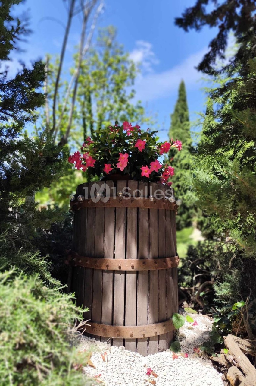 Un tonneau en bois transformé en jardinière avec des fleurs roses, entouré de verdure sous un ciel bleu.
