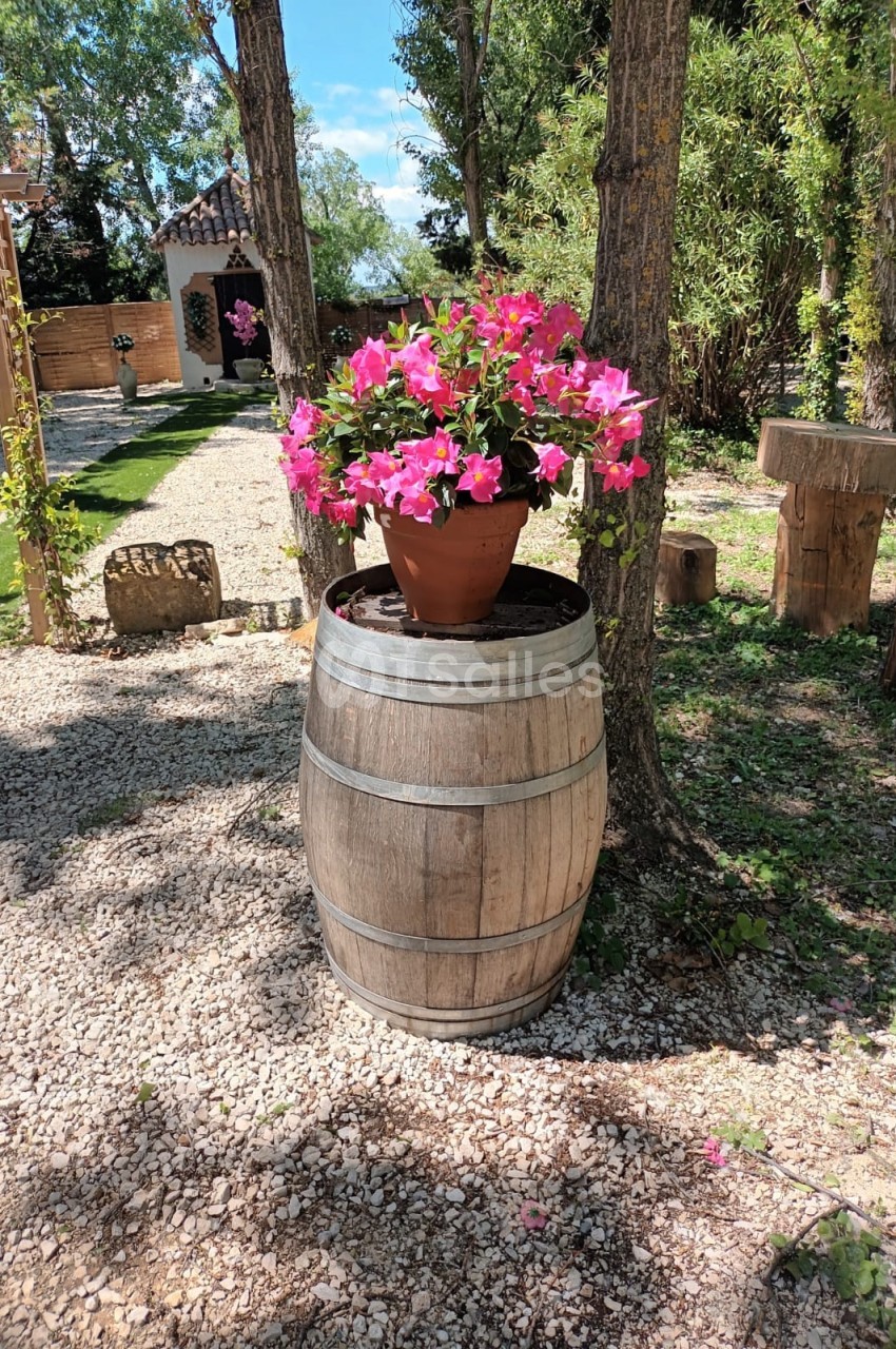 Pot de fleurs roses posé sur un tonneau en bois, dans un jardin ensoleillé entouré d'arbres et de mobilier rustique.