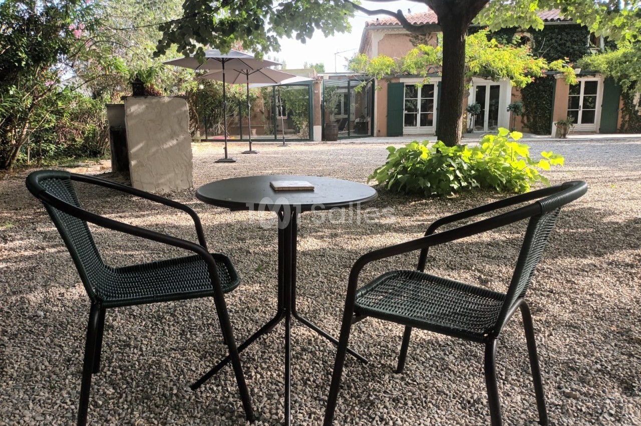 Deux chaises et une table ronde en métal noir sur un sol gravillonné, dans une cour ombragée par des arbres.