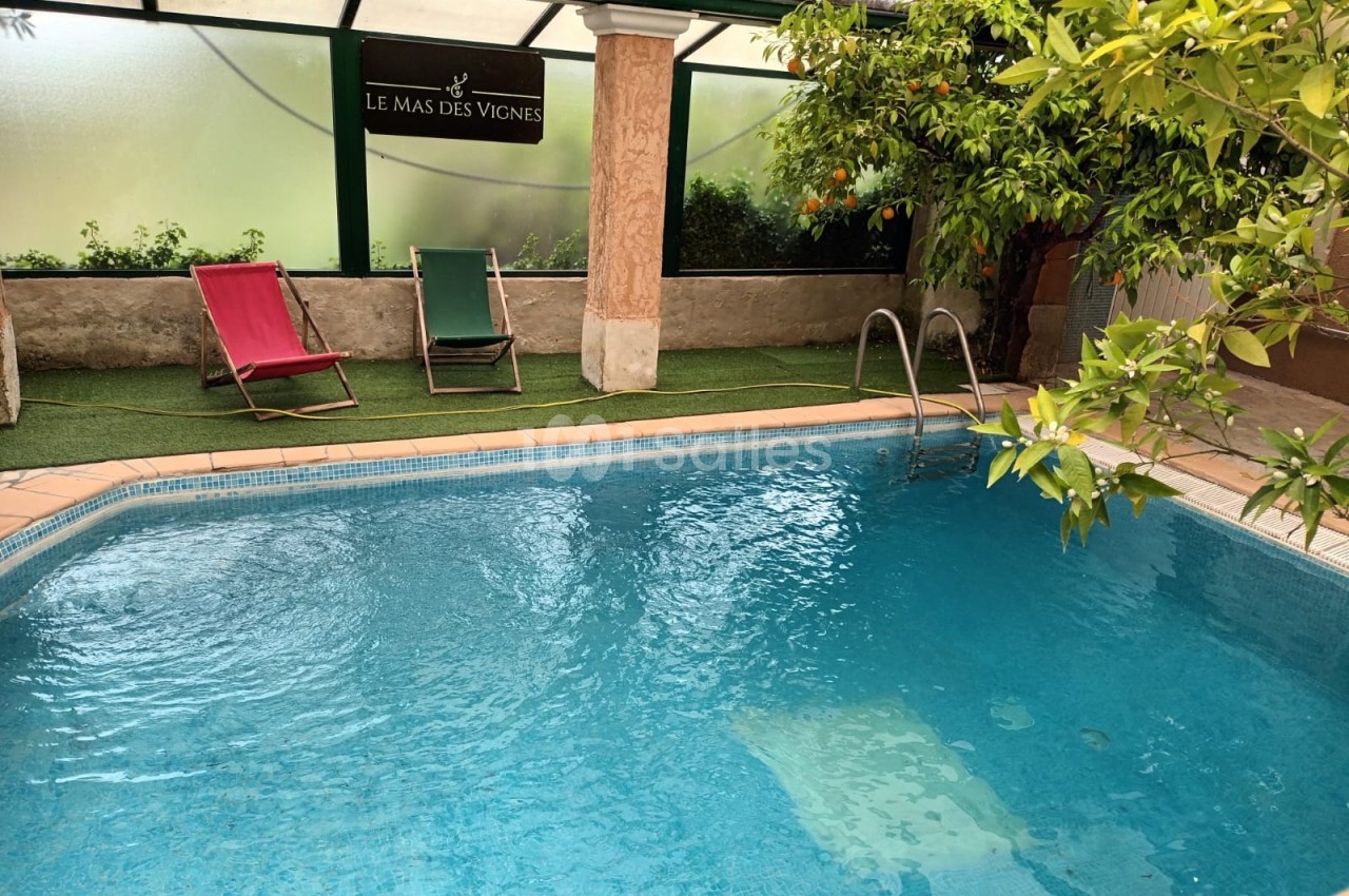 Piscine intérieure entourée de verdure avec deux chaises longues et un panneau indiquant ’Le Mas des Vignes’.