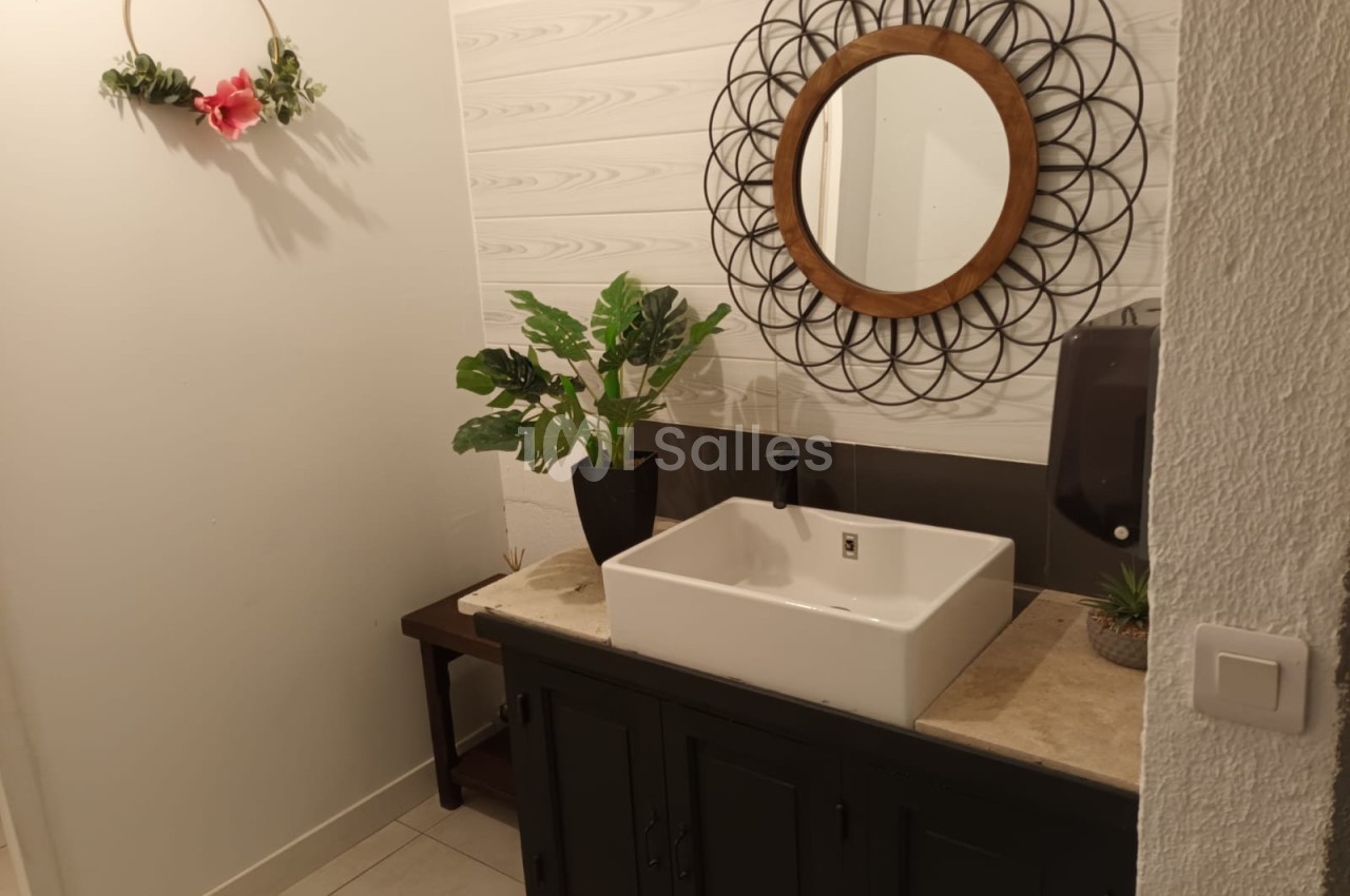 Salle de bain avec un lavabo blanc, un miroir rond encadré de métal, une plante verte et une décoration murale.