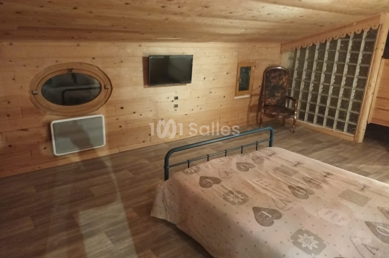 Chambre mansardée en bois avec lit double, télévision murale, fauteuil et cloison en briques de verre.