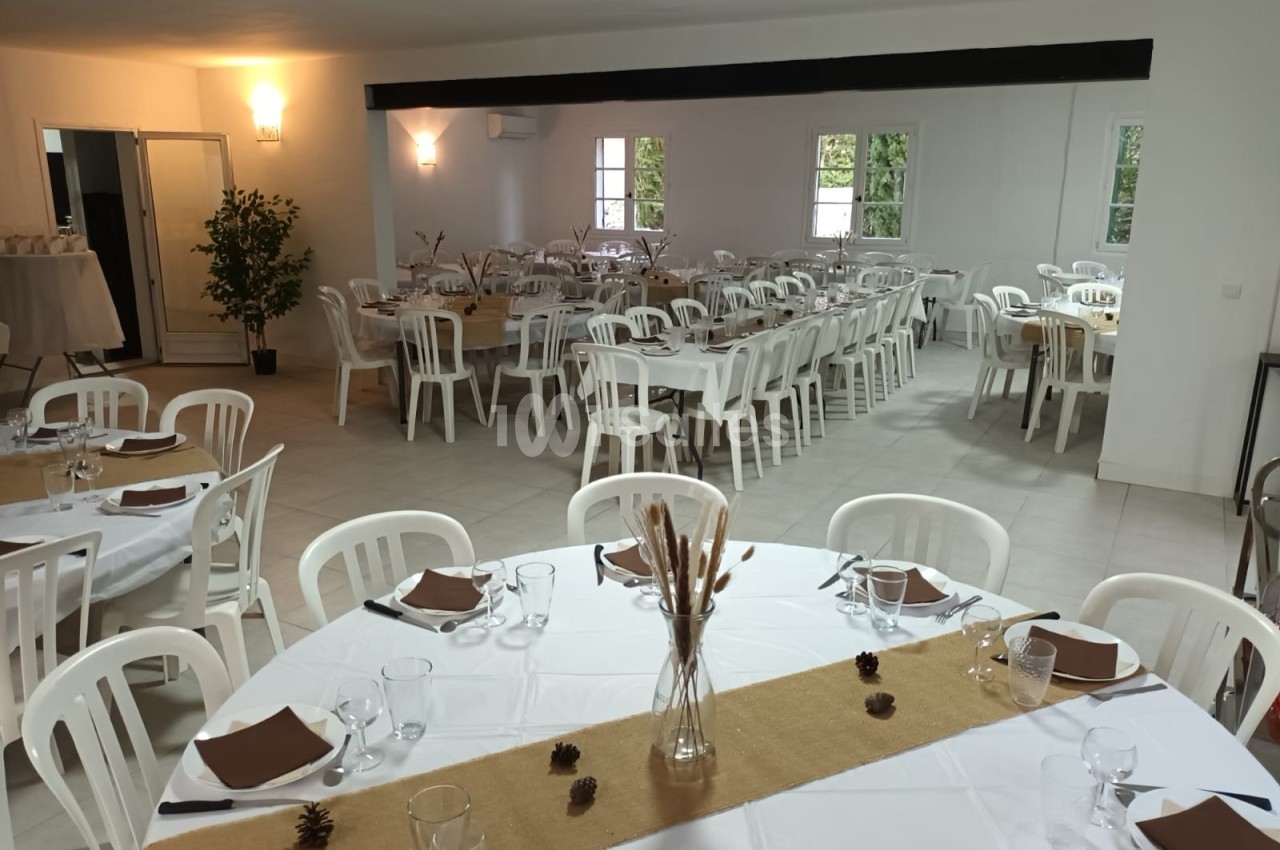Salle de réception lumineuse avec tables rondes et rectangulaires dressées, décorations simples et chaises blanches.