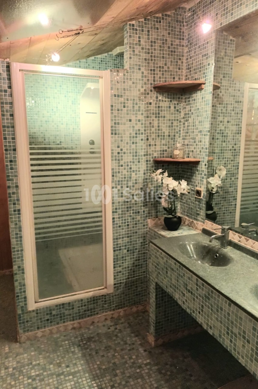 Salle de bain avec douche vitrée, murs en mosaïque bleue, double vasque et étagères murales en bois.