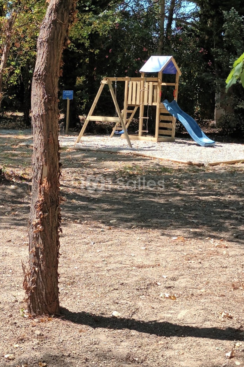 Structure de jeu en bois avec toboggan bleu et balançoires, située sur un sol gravillonné dans un espace extérieur arboré.