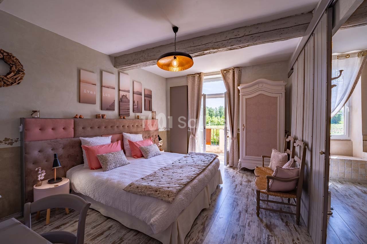 Chambre lumineuse avec lit double, décor pastel, armoire ancienne, et vue sur un paysage verdoyant par une porte-fenêtre.