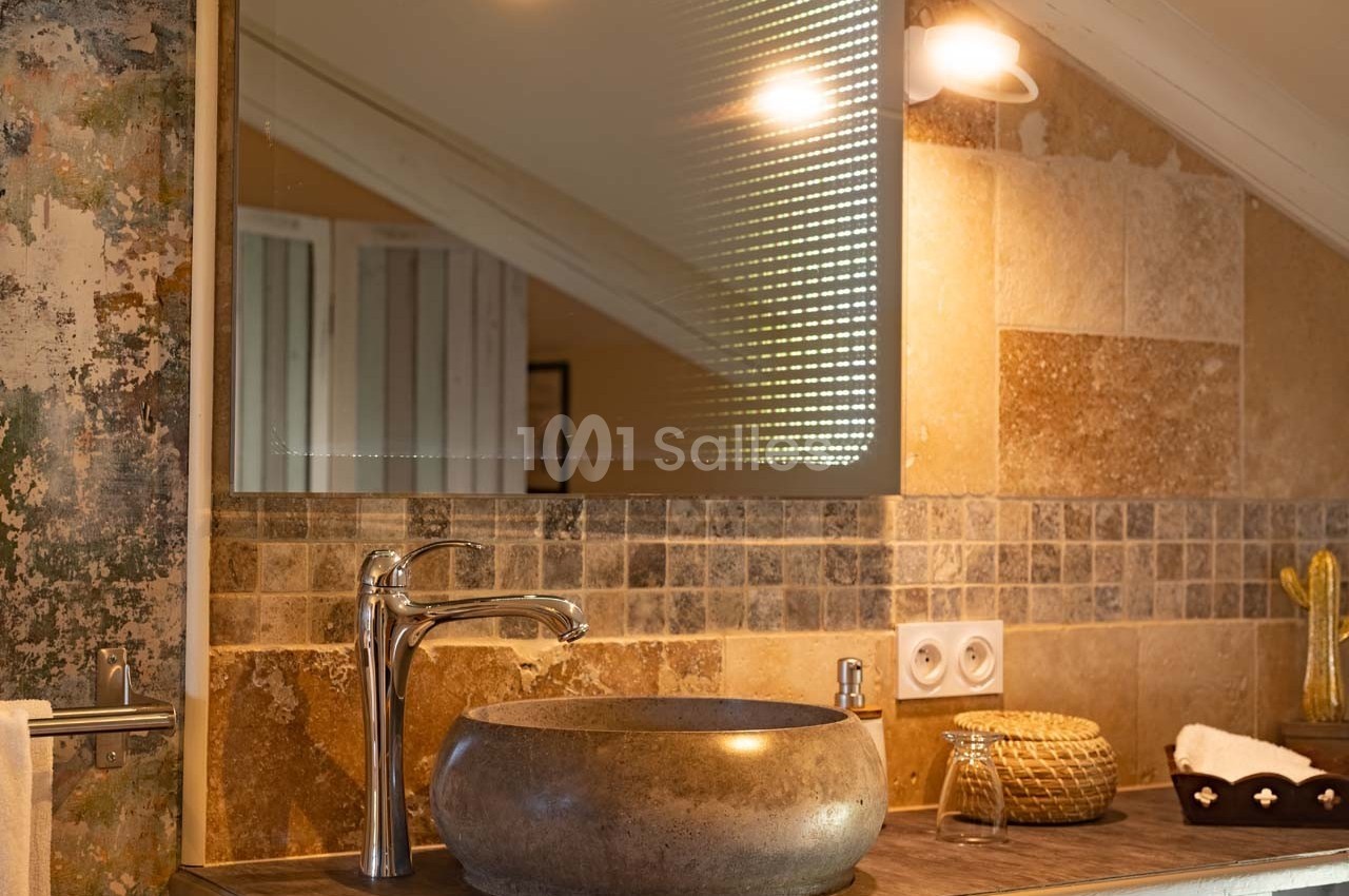 Lavabo en pierre sur un plan de travail en bois, avec miroir éclairé et carrelage mural beige dans une salle de bain.