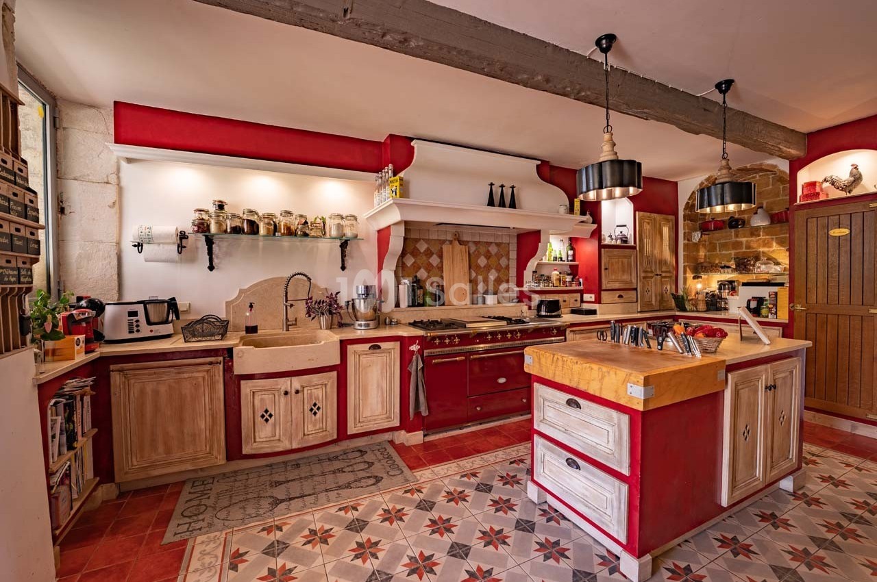 Cuisine rustique avec meubles en bois rouge et beige, plan de travail central, étagères ouvertes et carrelage décoratif.