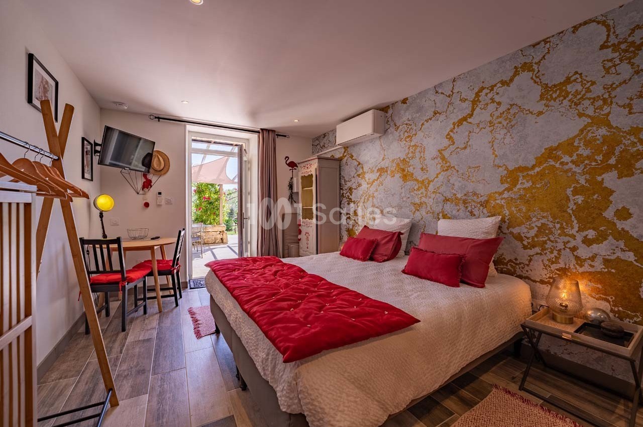 Chambre lumineuse avec lit double, décoration murale dorée, coin repas et accès à une terrasse extérieure.