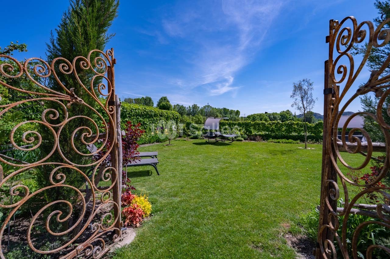 Portail en fer forgé ouvrant sur un jardin verdoyant avec pelouse, transats et ciel bleu dégagé.