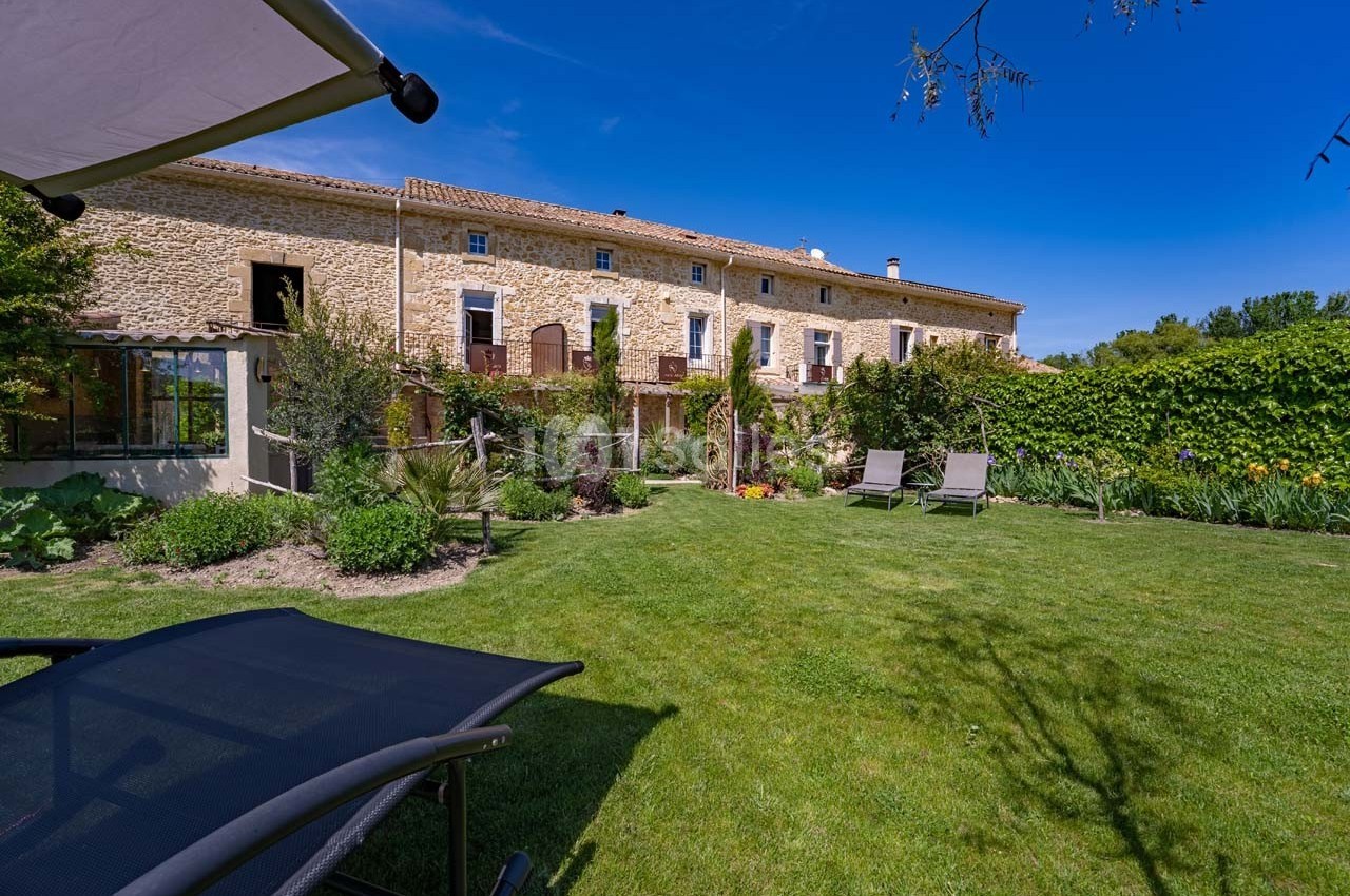 Façade en pierre d'une maison avec jardin verdoyant, chaises longues et parasol sous un ciel bleu.