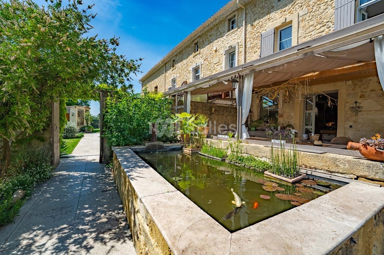 Bassin avec poissons et plantes aquatiques devant une maison en pierre avec terrasse couverte, entourée de verdure.