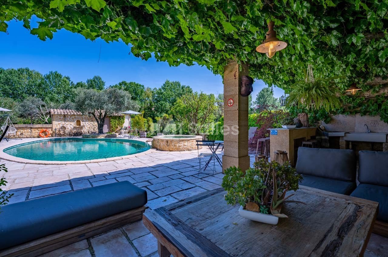 Terrasse ombragée avec mobilier en bois, vue sur une piscine entourée de végétation et d'oliviers.