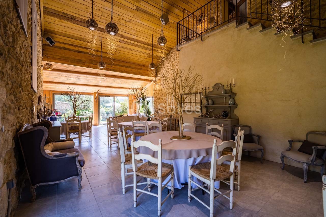 Salle lumineuse avec murs en pierre, plafond en bois, tables rondes dressées et grandes baies vitrées donnant sur un jardin.