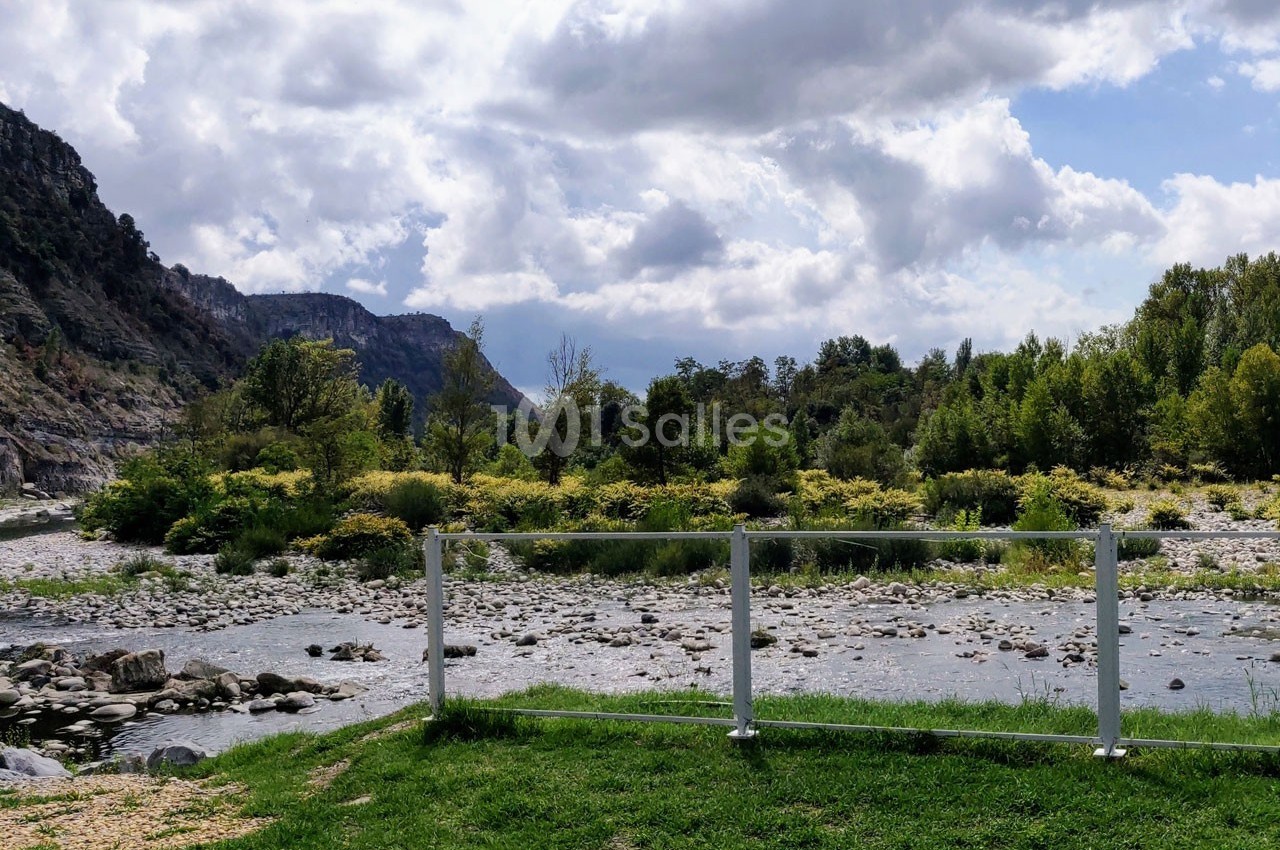 Rivière bordée de végétation et de montagnes sous un ciel partiellement nuageux, vue depuis une barrière blanche.