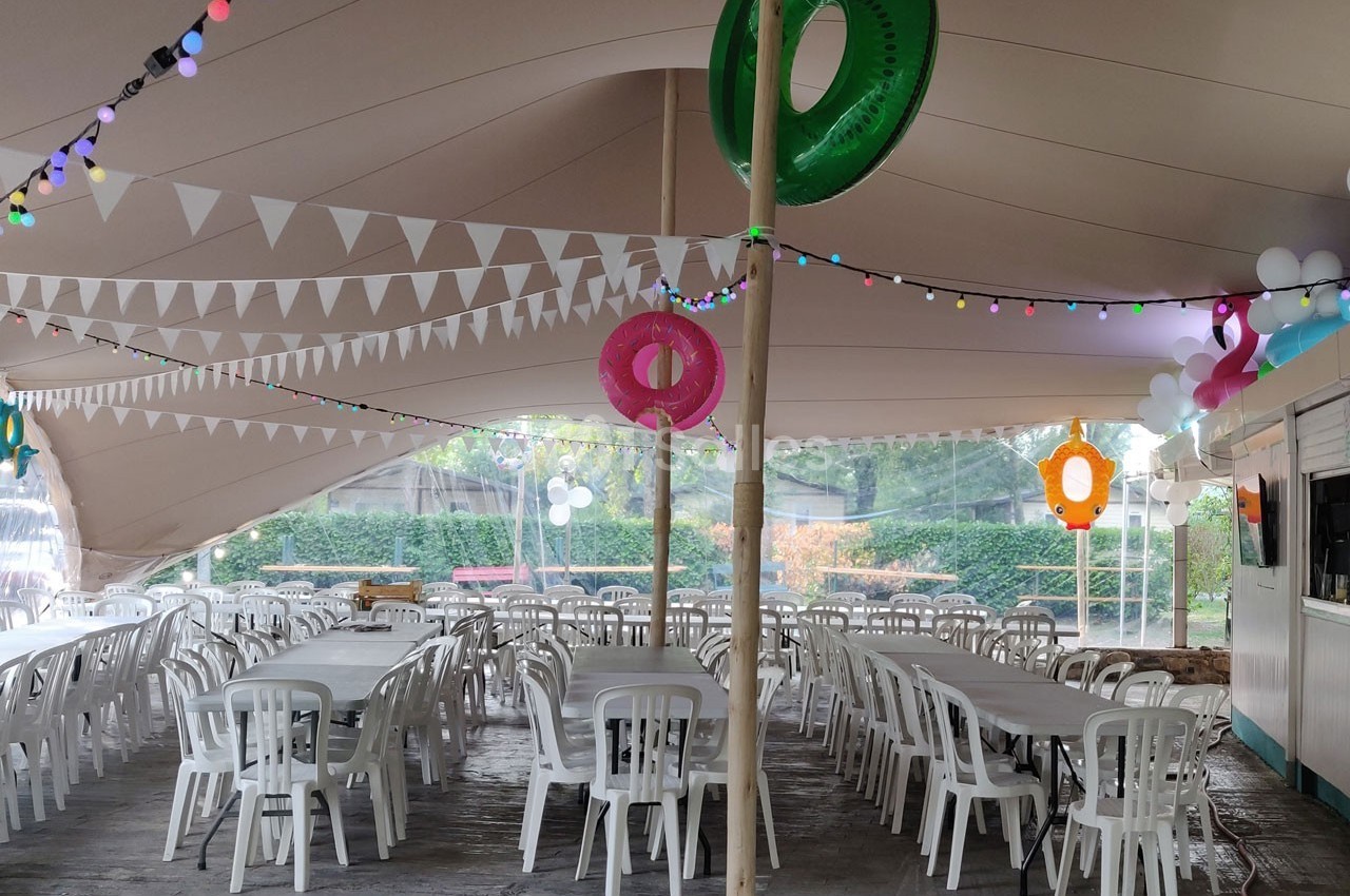 Intérieur d'une grande tente décorée avec des guirlandes lumineuses, des bouées colorées et des tables entourées de chaises…