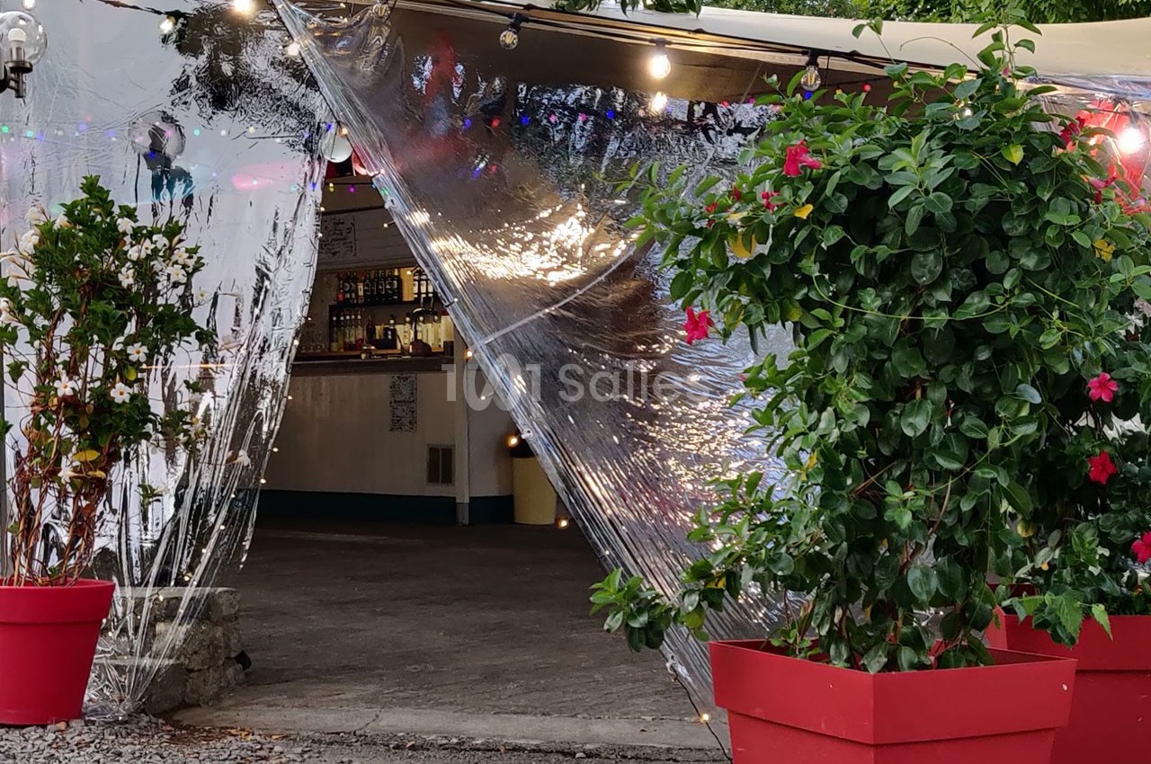 Entrée d'un espace extérieur couvert avec des plantes en pots rouges et une bâche transparente laissant entrevoir un bar.
